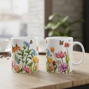 May include: Two white ceramic mugs with a floral design featuring colorful flowers, butterflies, and bees. The mugs are on a wooden surface, with a blurred background of a window and greenery.