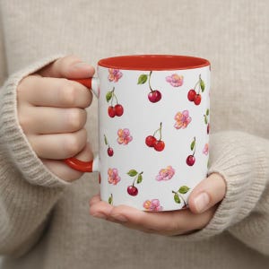 Könnte beinhalten: Ein weißer Keramikbecher mit einem wiederkehrenden Aquarellmuster aus roten Kirschen mit grünen Stielen und rosa Blüten. Der Becher hat ein durchgehend rotes Inneres und einen passenden roten Henkel, der einen Kontrast zur weißen Außenfläche bildet.
