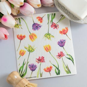 May include: A white, rectangular cleaning cloth with a watercolor floral design featuring tulips in shades of yellow, orange, red, and purple. Green stems and leaves are also present. The text "DICKSON.COM" is printed on the cloth.