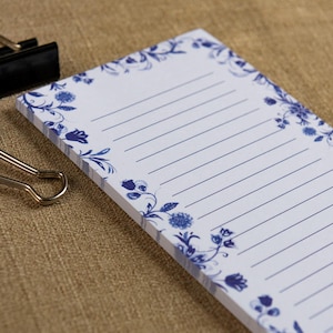 May include: A white notepad with blue floral corner designs and lined pages. The notepad is rectangular and has a metal clip on the side. The floral design is a classic blue and white pattern.