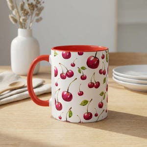 May include: A white ceramic mug featuring a repeating watercolor pattern of red cherries with green leaves. The mug has a bright red handle and a matching red interior rim, providing a vibrant contrast to the white exterior surface.