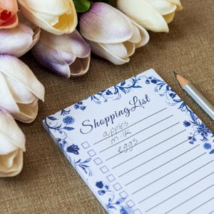 May include: A rectangular notepad with a blue floral border and the text Shopping List printed at the top. The list contains handwritten entries for apples, milk, and eggs next to checkboxes. A wooden pencil lies beside the notepad.