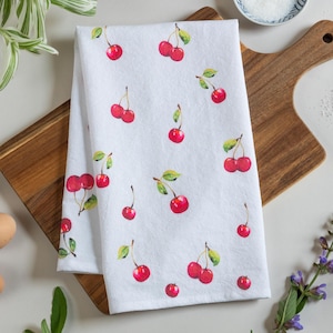 May include: A white kitchen towel featuring a repeating pattern of bright red cherries with green stems and leaves. The cherries are depicted in a watercolor style, scattered across the fabric in various groupings of single and paired fruits.