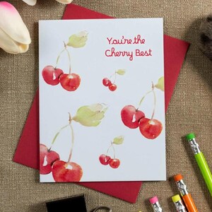 May include: A white ceramic mug with a red interior and handle, featuring a repeating pattern of small red cherries with green stems. A matching white greeting card displays watercolour cherries and the text You're the Cherry Best.