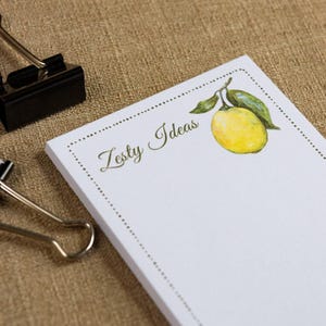 May include: White notepad with the text "Zesty Ideas" and a watercolor illustration of a lemon with green leaves. The notepad has a dotted border and is next to a black binder clip and a silver binder clip.