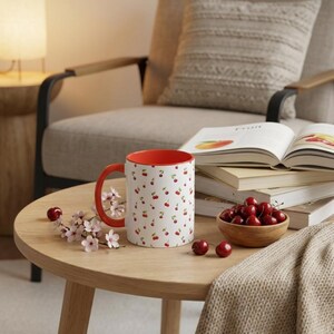 May include: A white ceramic mug with a repeating pattern of small red cherries and green stems, featuring a bright orange interior and handle. The mug is placed on a wooden surface alongside a small wooden bowl filled with fresh red cherries.