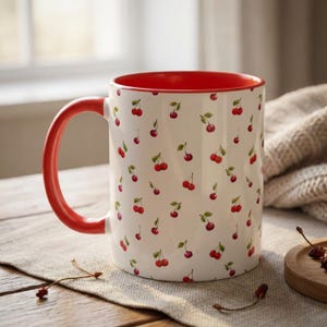 May include: A white ceramic mug featuring a repeating pattern of small red cherries with green stems. The mug has a bright red handle and a matching red interior, providing a vibrant contrast to the white exterior surface of the vessel.
