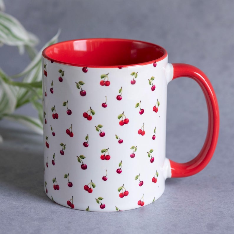 May include: A white ceramic mug featuring a repeating pattern of small red cherries with green stems. The interior of the mug and the curved handle are finished in a solid, vibrant red colour, providing a contrast to the white exterior surface.