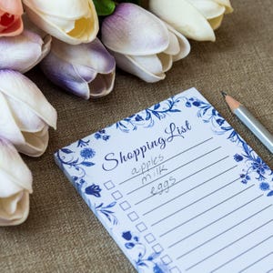 May include: A shopping list notepad with blue floral border and the words "Shopping List" in script. The list includes "apples, milk, eggs". A pencil rests on the side. Tulips in shades of white, purple, and pink are in the background.