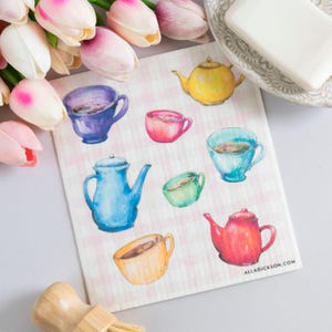 May include: A white and pink checkered tea towel featuring watercolor illustrations of teacups and teapots in various colors. The towel includes a blue teapot, a yellow teapot, and a red teapot. The text "ALLADICKSON.COM" is visible.