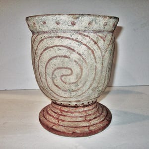 May include: A beige ceramic chalice featuring a wide, flared rim with a dotted pattern. The body is decorated with large, hand-painted reddish-brown spiral motifs. The base is circular and features concentric rings in the same reddish-brown color.