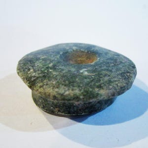 May include: A circular, mottled green stone object with a textured, speckled surface. The piece features a wide, flat top with a central circular indentation and a slightly narrower, cylindrical base, resembling a button or a small decorative knob.