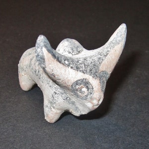 Puede incluir: Una pequeña figurita de toro de cerámica pintada a mano con marcas negras y blancas. El toro tiene una cara estilizada y cuernos grandes.