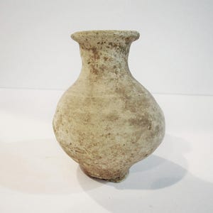 May include: A small, bulbous vase with a narrow neck and a flared rim. The surface of the vase is textured and matte, featuring a mottled, earthy beige and light brown color palette with visible signs of age and wear across its entire exterior.