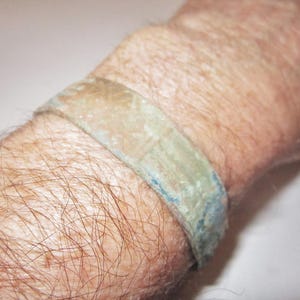 Könnte beinhalten: Nahaufnahme eines Handgelenks, das ein grün-blaues Armband trägt. Das Armband besteht aus einem strukturierten Material und wirkt alt oder abgenutzt.