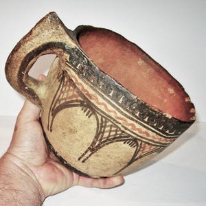 May include: An antique, hand-painted ceramic bowl with a handle. The bowl features a beige exterior with black and red geometric patterns. The interior is a reddish-brown color. The bowl is held in a hand.