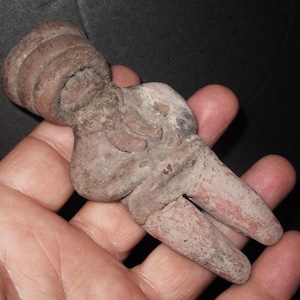 May include: A small, terracotta figurine of a person with a rounded head and a simple, stylized body. The figurine is hand-crafted and has a rustic, earthy appearance.