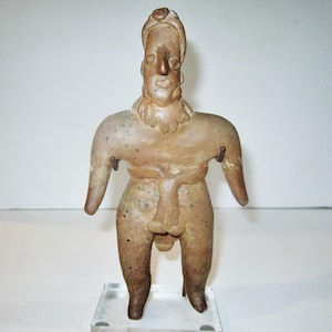 Pode incluir: Uma estatuinha de terracota de uma pessoa em pé com um chapéu. A estatuinha é marrom e tem um rosto detalhado com um nariz grande.