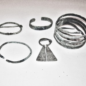 Könnte beinhalten: Eine Auswahl antiker Metallschmuckstücke, darunter eine Brosche, mehrere Armbänder und ein Anhänger. Die Artikel sind in Grau- und Brauntönen gehalten, mit sichtbaren Gebrauchsspuren, die auf Alter und Nutzung hindeuten. Der Schmuck ist auf einer weißen Oberfläche angeordnet.