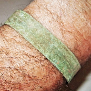 Könnte beinhalten: Ein rechteckiges Armband mit grüner Patina, getragen am Handgelenk. Das Metallband ist leicht gebogen. Die Oberfläche des Armbands weist Gebrauchsspuren auf. Das Handgelenk ist behaart.