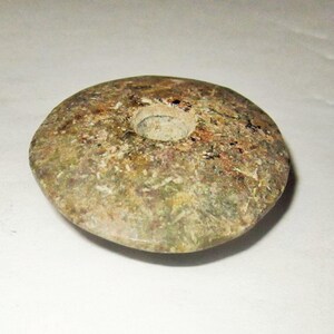 May include: A circular, flat stone object with a mottled, earthy appearance in shades of green, brown, and tan. The surface has a rough, natural texture, and there is a single, centered, circular hole drilled through the middle of the piece.