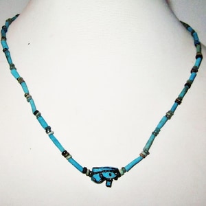 Puede incluir: Un collar de cuentas turquesas con un colgante del Ojo de Horus egipcio. El collar presenta pequeñas cuentas cilíndricas en tonos turquesa, acentuadas con cuentas más oscuras. El colgante es un símbolo de ojo estilizado en azul y negro.