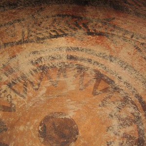 May include: Close-up of a brown earthenware pot with black painted designs. The designs include concentric circles, a sunburst, and a central circle.