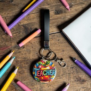 May include: A round keychain with a colorful apple design and the word "TEACHER" in blue, green, and yellow. The keychain has a black strap and silver hardware. Pencils and a notebook are scattered around the keychain on a wooden surface.