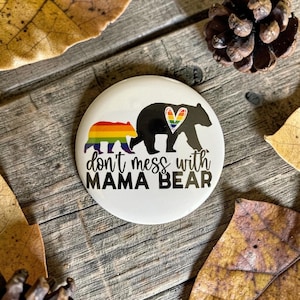 May include: A white button with a black bear silhouette and a rainbow bear cub. The text "don't mess with MAMA BEAR" is printed below the bears. The bear's heart is a rainbow design.