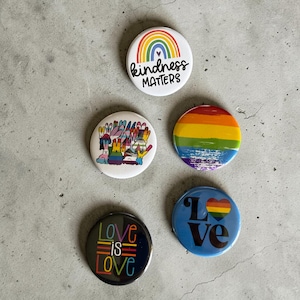 May include: A collection of five round pinback buttons with colorful designs. One button features a rainbow and the words "kindness matters." Other buttons display rainbow stripes, the words "Love is Love," and a heart.