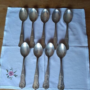 May include: Ten silver spoons with ornate designs on a white cloth background.