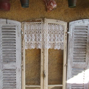 May include: A rustic scene featuring a white lace curtain over a double-door window with weathered, white-painted shutters. Above, a textured wall displays hanging metal buckets and a decorative pitcher, evoking a vintage, country aesthetic.