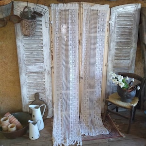 Può includere: Tende a crochet bianche con frange appese davanti a un set di persiane in legno bianco. Le persiane sono consumate e hanno un aspetto rustico. Le tende sono realizzate con un delicato motivo a pizzo. Le persiane sono parzialmente aperte, rivelando uno scorcio di un pavimento in legno.