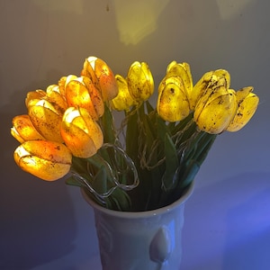 May include: A bouquet of illuminated yellow tulip lights in a white vase. The tulips have a warm, glowing effect, with green stems and leaves. The vase has a decorative design and is held by a hand.