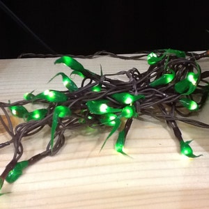 May include: A string of green LED fairy lights with dark brown wiring. The lights are clustered together, with each light emitting a bright green glow. The lights are designed to resemble small, stylized leaves.
