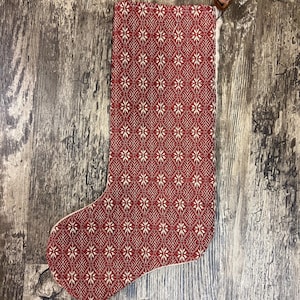 May include: A red and cream Christmas stocking with a geometric pattern. The stocking has a cream-colored interior and a wooden loop for hanging. The pattern is a repeating diamond shape with floral-like details.