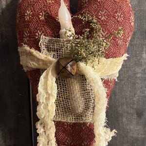 May include: A heart-shaped, red fabric decoration with a cream-colored candle and a burlap pocket. The heart is adorned with a cream-colored ribbon and a small wooden bell. The fabric has a subtle floral pattern.