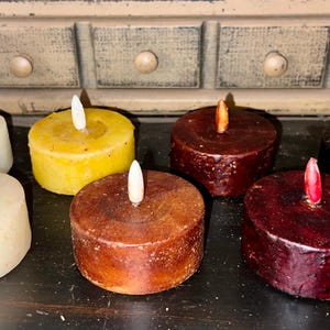 May include: Six unscented tea light candles in various colors, including white, yellow, brown, and red. The candles are arranged in a row on a dark surface.