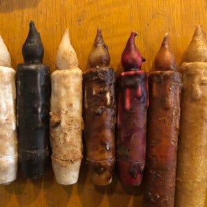 May include: A collection of seven antique-style candles in varying colors. The candles range from white to black, with shades of brown and red. Each candle has a textured, aged appearance, with a flame-shaped top.