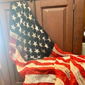 May include: An American flag with red and white stripes and a blue field of white stars. The flag is draped over a dark brown wooden cabinet. The flag appears to be vintage or antique.