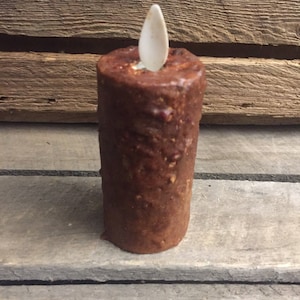 May include: A brown, battery-operated candle with a textured surface and a white, flame-shaped LED. The candle is cylindrical and sits on a wooden surface, evoking a rustic aesthetic.