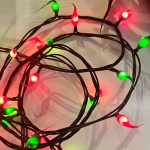 May include: A string of decorative holiday lights featuring dark brown wiring and flame-shaped bulbs in alternating red and green colors. The bulbs are illuminated, casting a soft glow, and are arranged in a tangled, circular coil pattern.