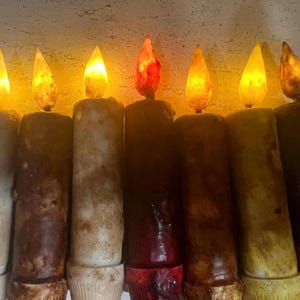 May include: A set of eight faux candles with a warm glow. The candles are in various shades of brown, beige, and red. The candles are lit and have a realistic flame effect.
