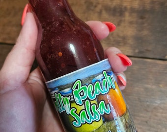Può includere: Una bottiglia di vetro di salsa rossa con un'etichetta che recita "Salty Beach Salsa" e include le informazioni di contatto: 830-998-9693, 1712 Hardee St., Rockport, Texas 78382.