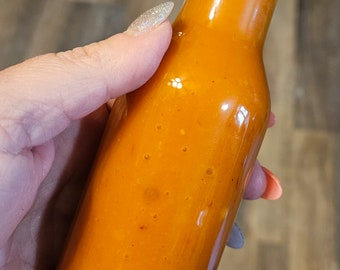 Può includere: Una bottiglia di vetro di salsa piccante arancione con un tappo nero. La bottiglia è piena di salsa e si vedono alcune bolle d'aria.