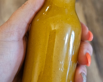 Può includere: Una bottiglia di vetro con un tappo nero riempita di un liquido giallo. La bottiglia è tenuta da una mano.