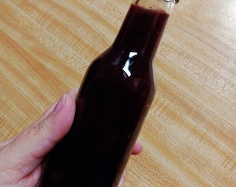 Può includere: Una bottiglia di vetro con un tappo nero riempita di un liquido rosso scuro. La bottiglia è tenuta in una mano.