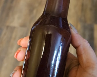 Può includere: Una bottiglia di vetro con un tappo nero riempita di un liquido viola scuro. La bottiglia è tenuta da una mano.