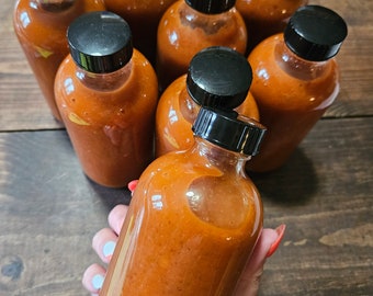 May include: A group of eight glass bottles filled with a red-orange liquid. The bottles have black screw-on lids and are arranged in a circle on a wooden surface.