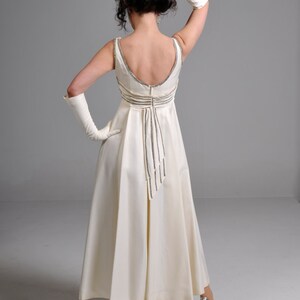 May include: A white floor-length gown with a low back and a beaded detail down the back. The gown is worn by a person with dark hair and white gloves. The person is wearing silver heels.